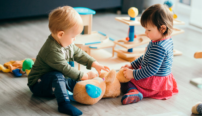 Foto: Kleiner Junge und kleines Mädchen sitzen am Fußboden und spielen mit einem Teddybär