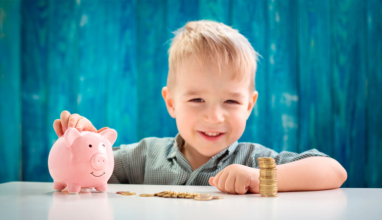 Foto: kleiner Junge sitzt mit seinem Sparschwein und vielen Geldmünzen lachend an einem Tisch