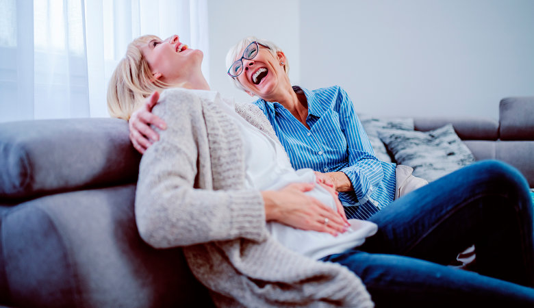 Foto: Schwangere Frau sitzt mit ihrer Mutter auf dem Sofa und lachen zusammen.