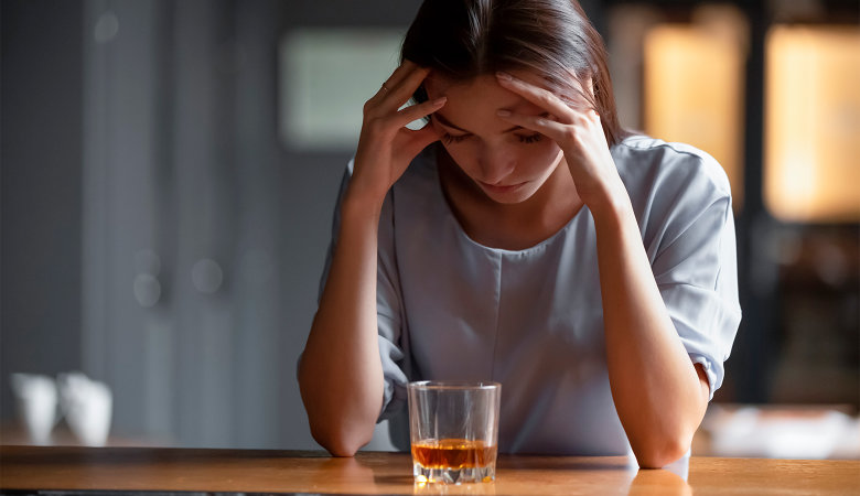 Foto: Depressive, traurige Frau sitzt am Tisch und hat ein Glas Alkohol vor sich auf dem Tisch stehen.