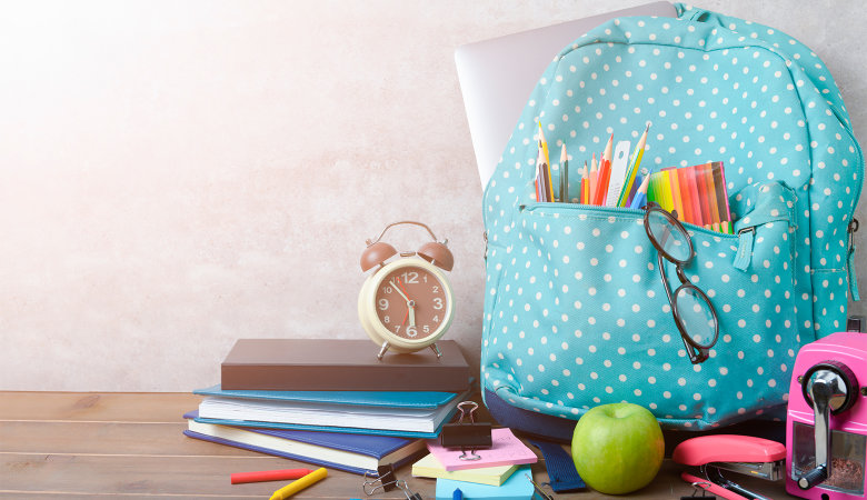 Foto: Schulrucksack, Notizbücher, Buntstifte, Apfel, Spitzer und Tacker liegen auf einem Holzboden