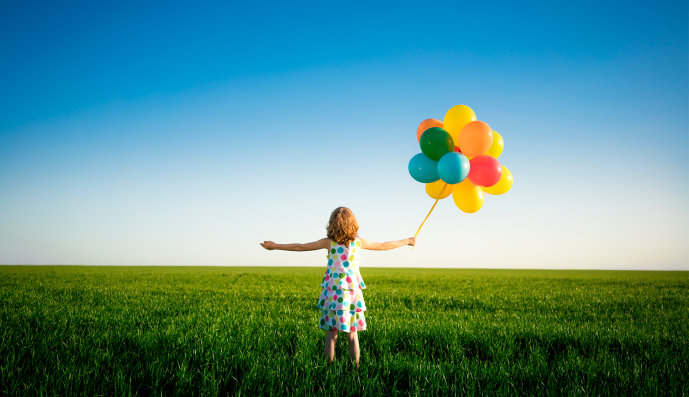 Foto: Mädchen steht mit ausgebreiteten Armen auf einer Wiese und hat viele Luftballons in der rechten Hand.