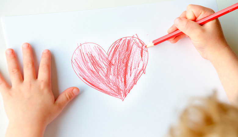 Foto: Kind zeichnet mit  einem Holzmalstift ein Herz auf ein Blatt Papier