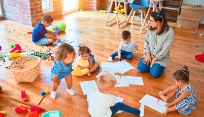 Foto: Kindergärtnerin sitzt mit sechs Kindern auf den Fußboden beim Spielen und Malen