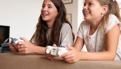 zwei Mädchen sitzen am Tisch und haben je eine Spielekonsole in der Hand