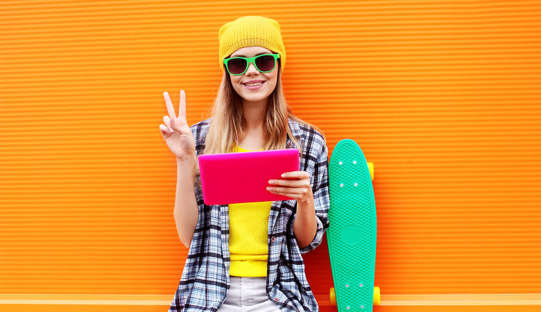 Foto: Junges Mädchen mit Mütze und Sonnenbrille lehnt mit einem Tablet in der Hand an einer Wand.