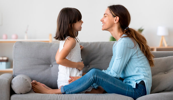 Foto: Lachende Mutter und ihre kleine Tochter sitzen sich auf dem Sofa gegenüber und sprechen miteinander.