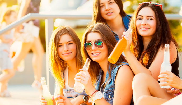 Foto: Vier junge Mädchen, die im Freien zusammen sitzen und alkoholische Mixgetränke aus der Flasche trinken.