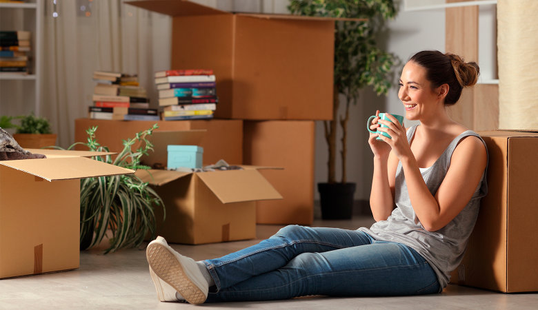 Foto: Junges Mädchen sitzt lachend auf dem Fußboden in ihrer Wohnung mit gepackten Umzugskartons.