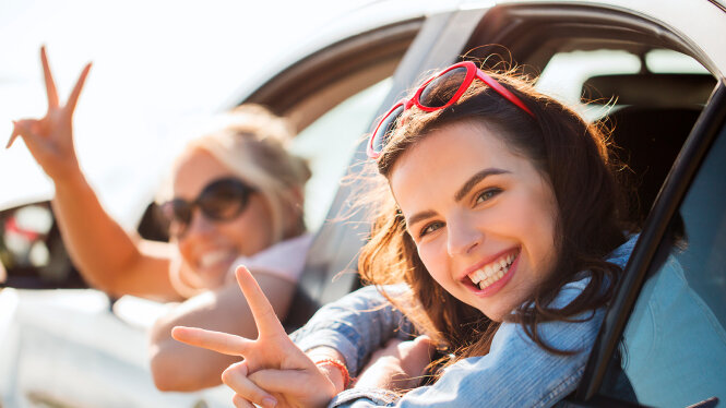 Foto: Zwei lachende Teenagermädchen lehnen sich aus dem Autofenster und winken.