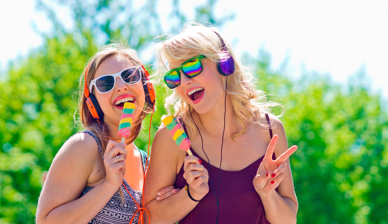 Foto: Zwei singende und lachende Teenagerinnen, die Kopfhörer und Sonnenbrillen aufhaben.