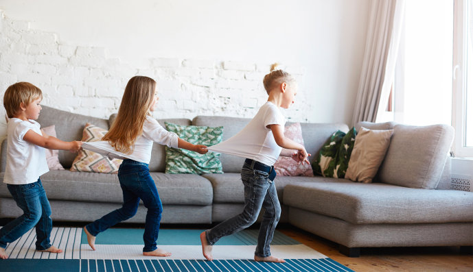 Foto: Drei Kinder tanzen hintereinander im Wohnzimmer und ziehen sich an den T-shirts