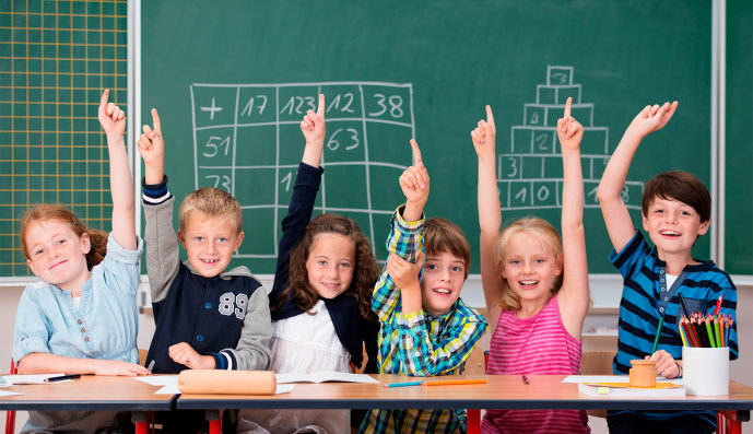 Foto: Begeisterte Gruppe von fünf jungen Kindern im Unterricht, die in einer Reihe am Schreibtisch sitzen und die Hände in die Luft heben