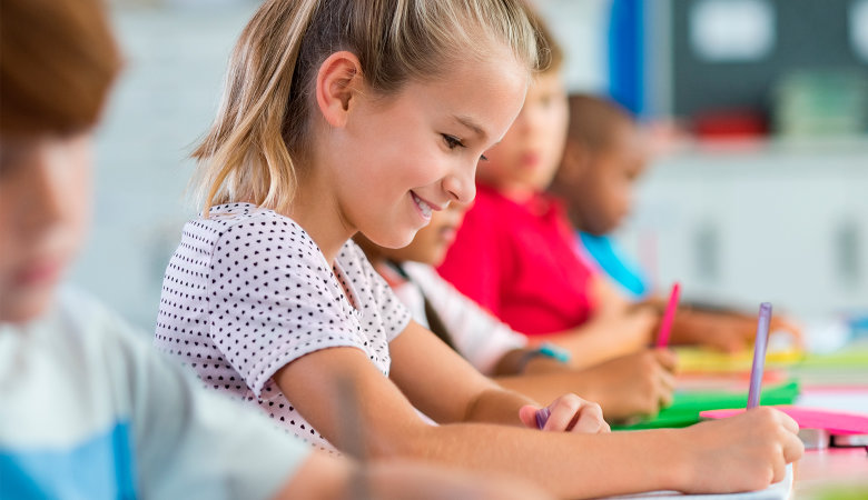 Foto: Lächelndes Mädchen sitzt im Klassenzimmer an ihrem Schreibtisch und schreibt.